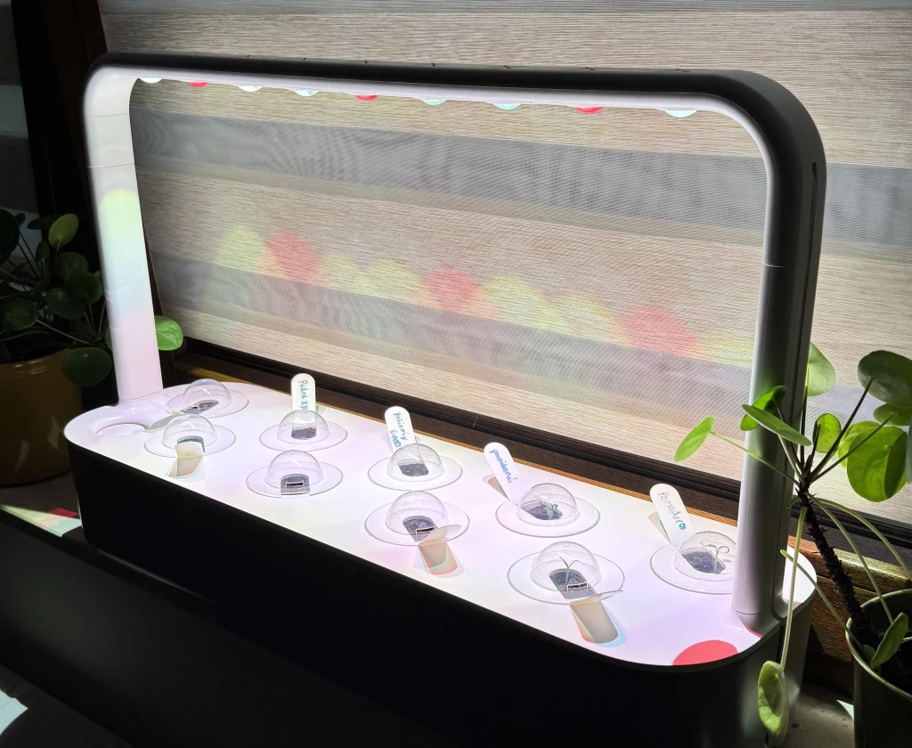 Nowoczesny zestaw do domowej uprawy roślin z podświetleniem LED, w którym znajdują się specjalne pojemniki z nasionami. Po obu stronach widoczne są rośliny doniczkowe ustawione na parapecie okiennym.