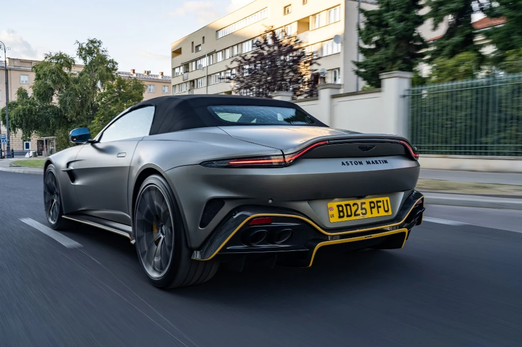 Matowy kabriolet Aston Martin z czarnym dachem i żółtymi akcentami na tyle pojazdu jedzie miejską ulicą, w tle nowoczesna zabudowa i drzewa.