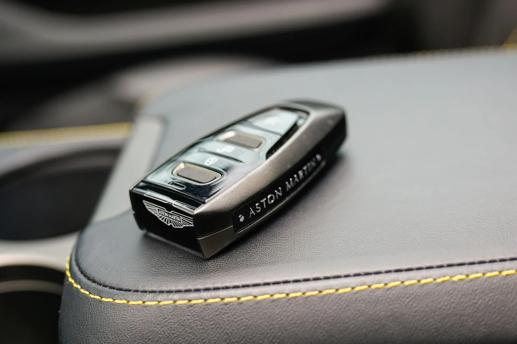 Czarny pilot do samochodu marki Aston Martin leżący na ciemnym skórzanym podłokietniku z żółtym przeszyciem.