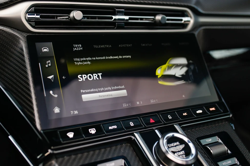 Ekran dotykowy samochodu wyświetlający menu ustawień trybu jazdy z zaznaczoną opcją SPORT, otoczony elementami konsoli środkowej oraz przyciskami do sterowania funkcjami pojazdu.
