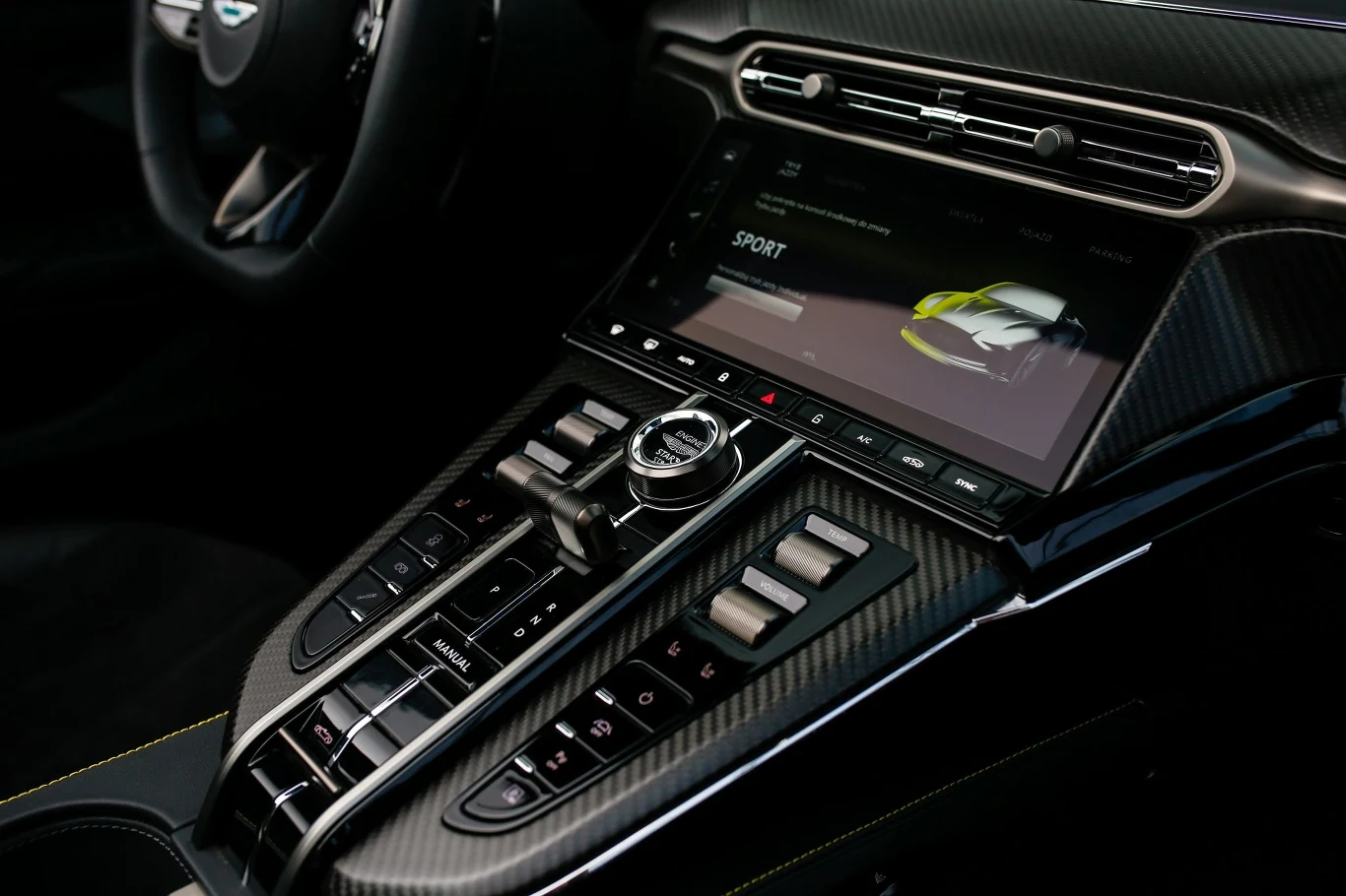 Za pomocą fizycznych przełączników można sterować nagłośnieniem czy regulować pracę zawieszenia. Nowoczesna konsola środkowa luksusowego samochodu z dużym ekranem dotykowym wyświetlającym ustawienia trybu jazdy, otoczona przyciskami sterującymi oraz elementami z włókna węglowego.