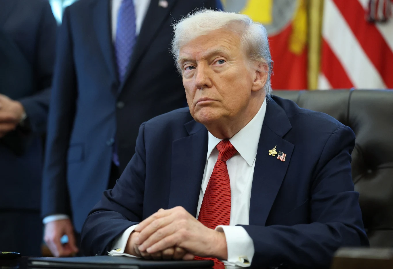 Administracja Donalda Trumpa inwestuje w firmę eksploatującą bogactwa mineralne Alaski Mężczyzna w garniturze siedzący za biurkiem, w tle obecni inni ludzie w formalnych strojach, widoczne amerykańskie flagi i złota odznaka na klapie marynarki.