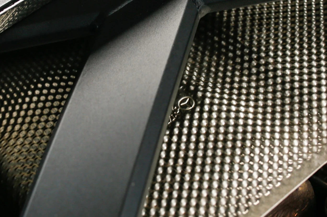 W komorze silnika można znaleźć oznaczenie Mercedesa. Metalowa powierzchnia z wypukłymi okrągłymi wzorami oraz element konstrukcyjny w kształcie litery X wykonany z czarnego materiału, razem tworzący geometryczną kompozycję z wyraźną fakturą.