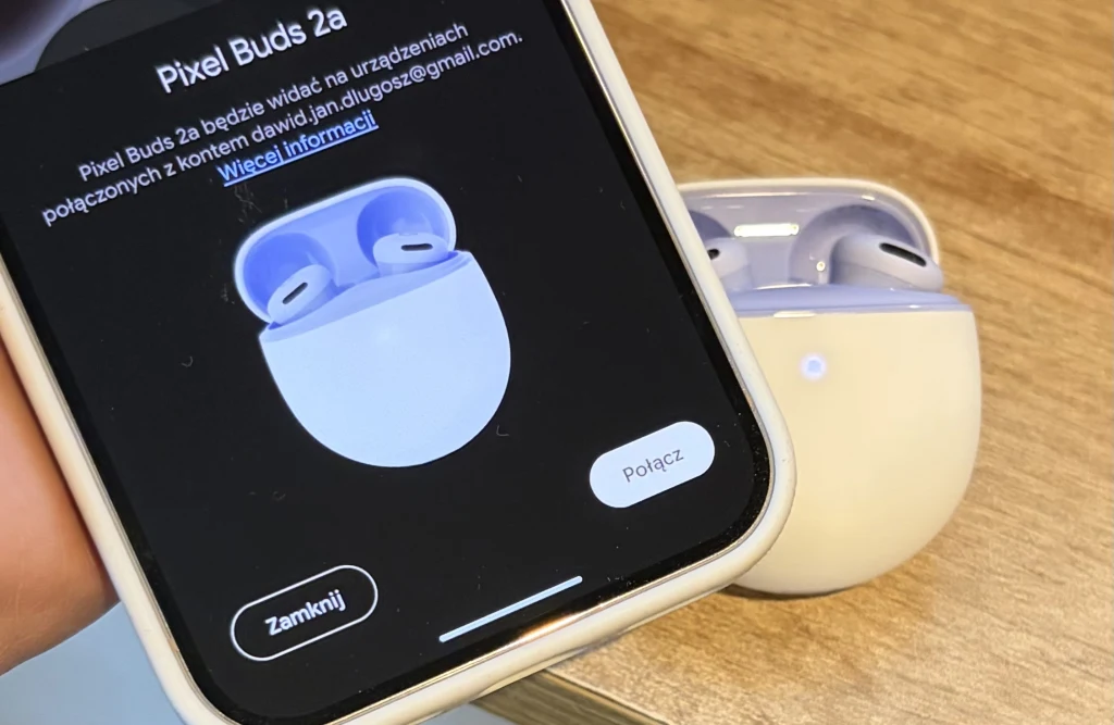 Białe słuchawki douszne Pixel Buds 2a w etui ładującym na drewnianym blacie oraz ekran telefonu z interfejsem parowania tych słuchawek.