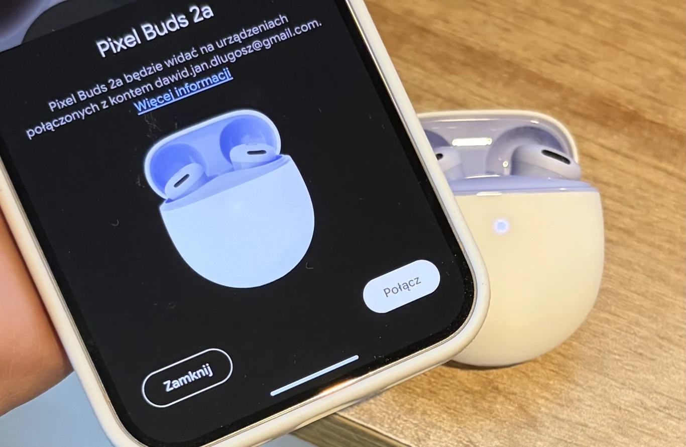 Białe słuchawki douszne Pixel Buds 2a w etui ładującym na drewnianym blacie oraz ekran telefonu z interfejsem parowania tych słuchawek.