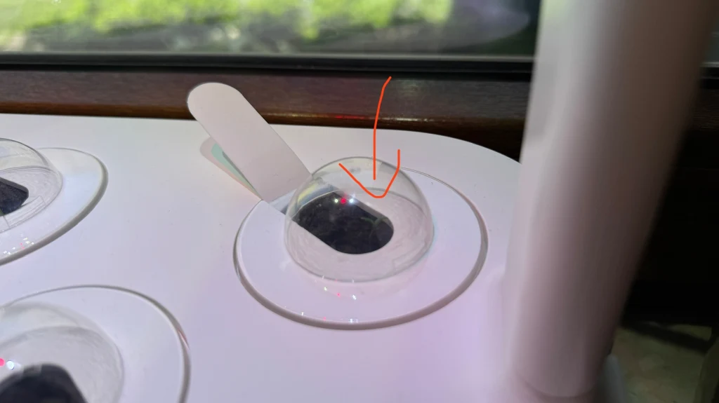 Pod przezroczystą kopułą w pojemniku z hydroponicznym systemem do uprawy roślin znajduje się nasiono lub pellet torfowy, a obok wystaje biały znacznik do ewentualnego opisu rośliny.