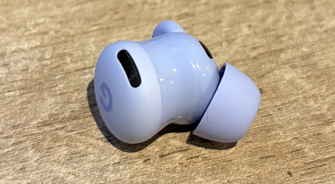 Jasnofioletowa bezprzewodowa słuchawka douszna leży na jasnym drewnianym blacie, widoczne logo na obudowie oraz otwory do emisji dźwięku i mikrofonu.