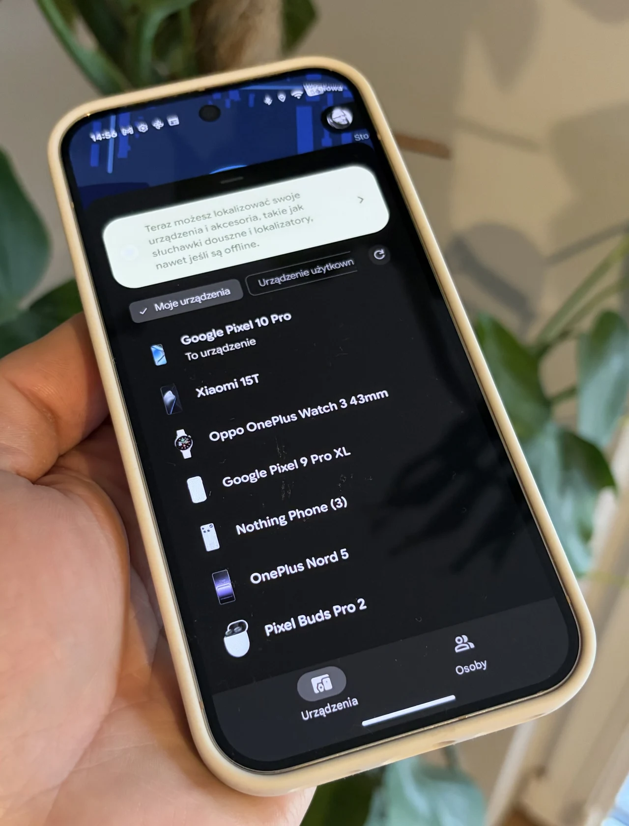 Pierwszy plan stanowi dłoń trzymająca smartfon z uruchomioną aplikacją wyświetlającą listę różnych urządzeń elektronicznych takich jak smartwatche, telefony i słuchawki bezprzewodowe, tło rozmyte z widoczną rośliną domową.