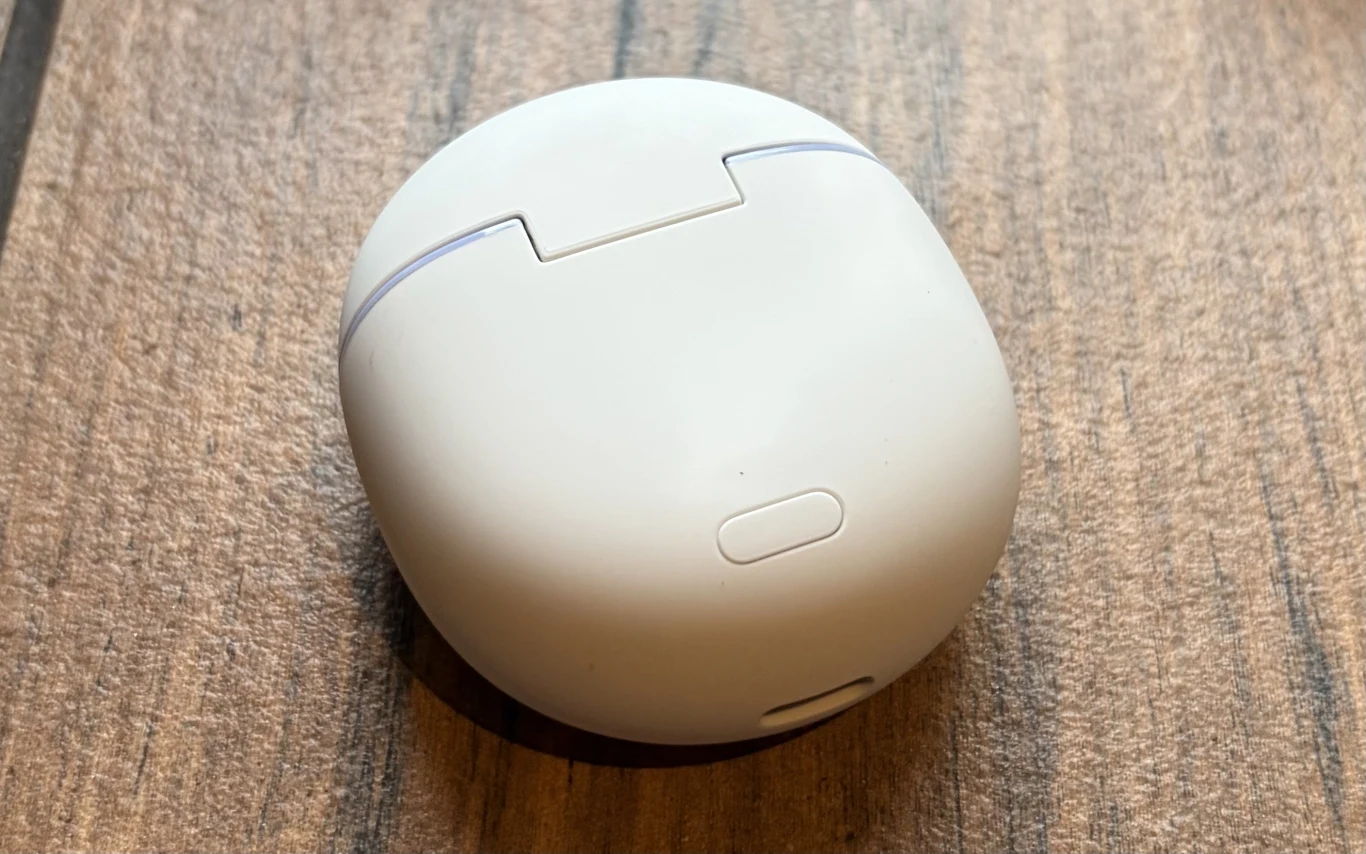 Białe, zaokrąglone etui na słuchawki bezprzewodowe leży na drewnianej powierzchni; widoczny jest zatrzask i przycisk ułatwiający otwieranie futerału.