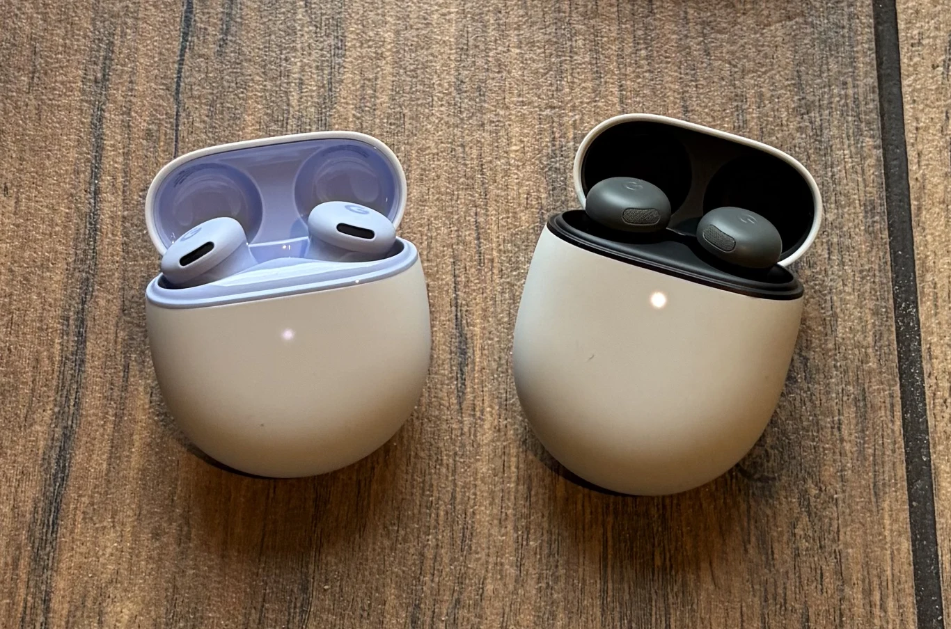 Dwie pary bezprzewodowych słuchawek dousznych w otwartych etui ładujących ustawionych na drewnianym blacie, jedna para w jasnym etui, druga w ciemniejszym wnętrzu.
