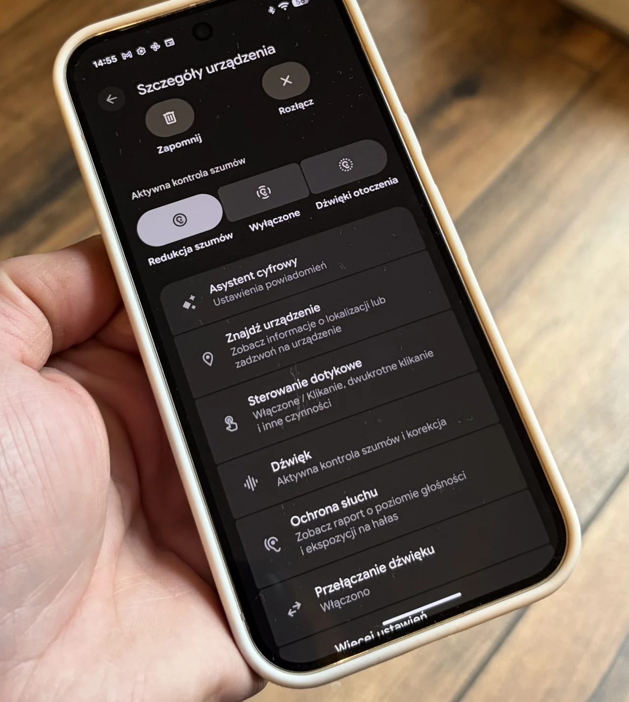 Smartfon w jasnym etui trzymany w lewej dłoni, na ekranie widoczne są opcje ustawień dotyczących szczegółów urządzenia, takich jak aktywna kontrola szumów, asystent cyfrowy, sterowanie dotykowe oraz ochrona słuchu, tło stanowią drewniane panele podłogowe.