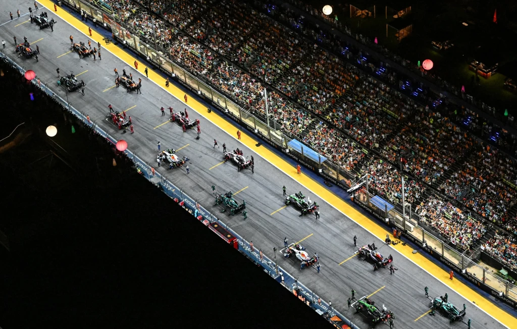 Tor wyścigowy oświetlony nocą z ustawionymi bolidami Formuły 1 oraz zespołami technicznymi, po bokach trybuny wypełnione kibicami.