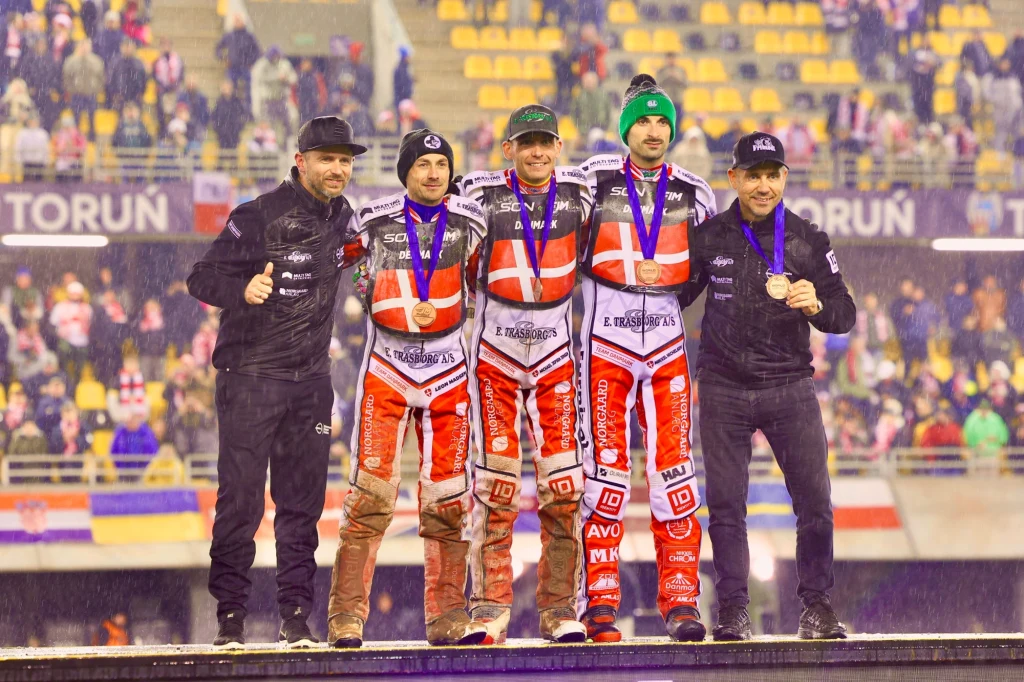 Reprezentacja Danii z brązowym medalem SoN 2025 Czterech żużlowców w biało-czerwonych kombinezonach z medalami oraz dwóch trenerów stoi na podeście stadionu podczas ceremonii dekoracji, w tle trybuny z kibicami.
