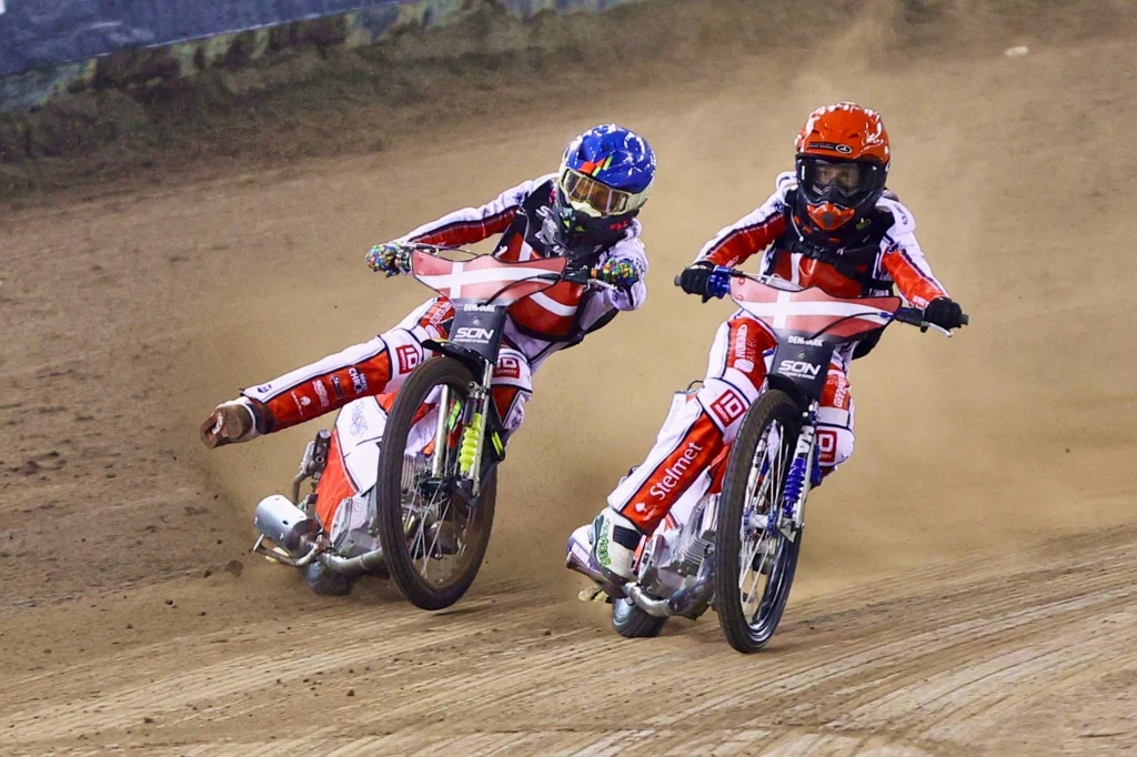 Madsen i Jensen w akcji podczas SoN w Toruniu Dwóch żużlowców w czerwono-białych kombinezonach rywalizuje na torze żużlowym, jadąc na motocyklach przez zakręt w chmurze pyłu.