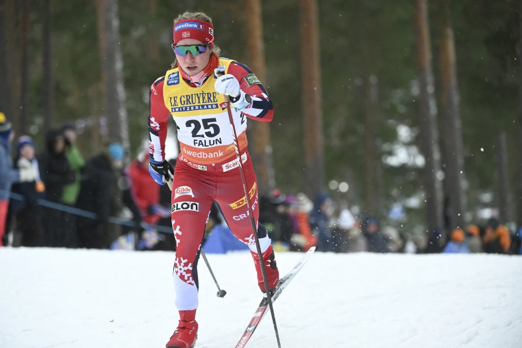 Narciarz biegowy w czerwonym stroju z numerem 25 podczas zawodów, dynamicznie poruszający się na tle ośnieżonej trasy i widzów oraz drzew w tle.