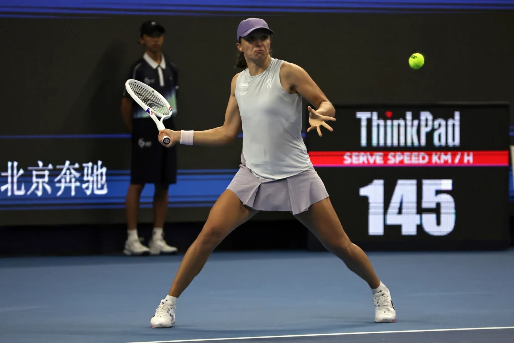 Iga Świątek tenisistka w fioletowej spódniczce i czapce przygotowuje się do uderzenia piłki na korcie tenisowym, w tle widoczny sędzia przy linii oraz elektroniczny licznik prędkości serwisu wyświetlający liczbę 145