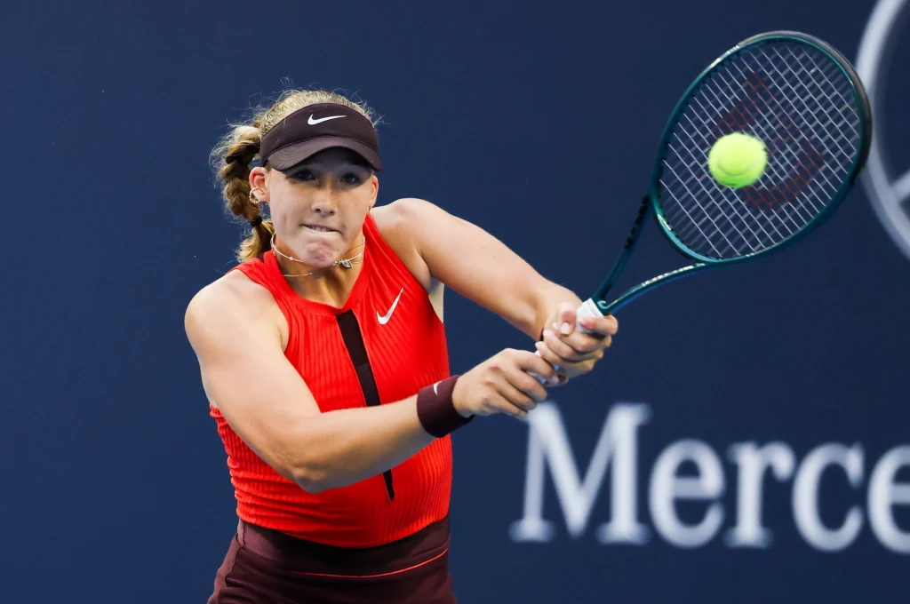 Mirra Andriejewa Zawodniczka w czerwonym stroju sportowym uderza piłkę tenisową rakietą podczas meczu, skupiona na dynamicznej akcji na kortcie.