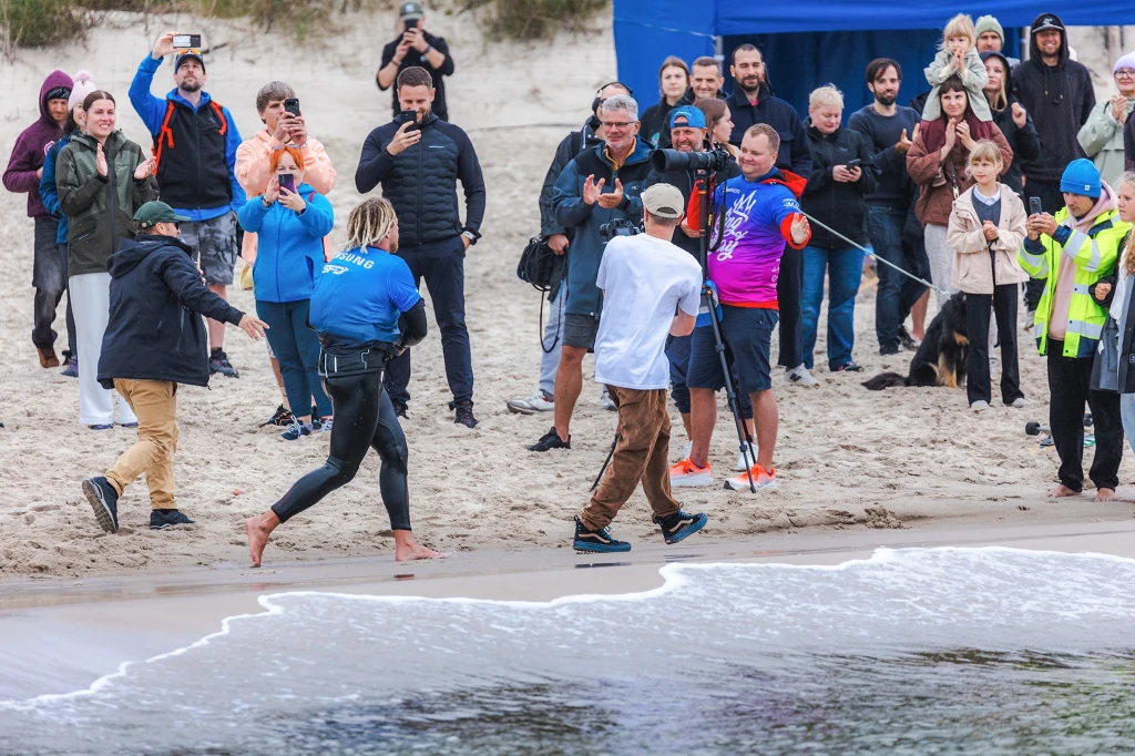 Maciek Rutkowski wbiega na metę wrześniowego wyzwania Tłum ludzi zgromadzony na plaży, część z nich fotografuje i filmuje osobę w stroju sportowym oraz inną osobę, która trzyma mikrofon; wokół obecni są widzowie i osoby organizujące wydarzenie.