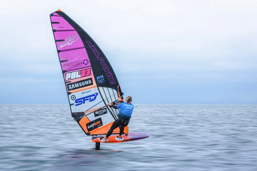 Maciek Rutkowski podczas Misji Bałtyk Sportowiec ubrany w niebieski strój pływa na pomarańczowo-fioletowej desce windsurfingowej po spokojnej tafli wody, trzymając się dużego żagla ozdobionego licznymi naklejkami sponsorów.