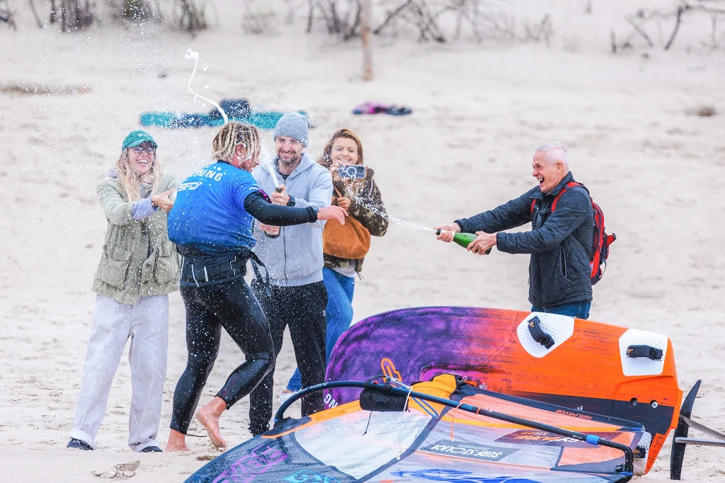 Po przekroczeniu mety rozpoczęło się prawdziwe świętowanie Grupa osób celebruje sukces na plaży, jedna osoba polewa inną szampanem w otoczeniu deski windsurfingowej, wszyscy są uśmiechnięci i zadowoleni