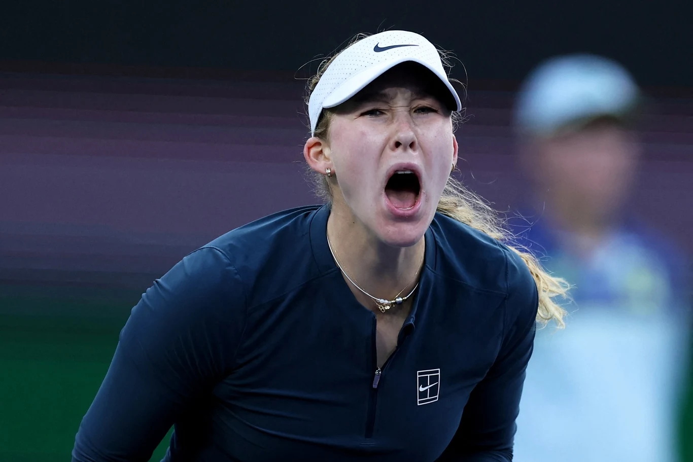 Tenisistka w sportowym stroju i białej czapce z daszkiem wyraża silne emocje podczas meczu, jej usta są szeroko otwarte, a twarz wyraża intensywność przeżycia.