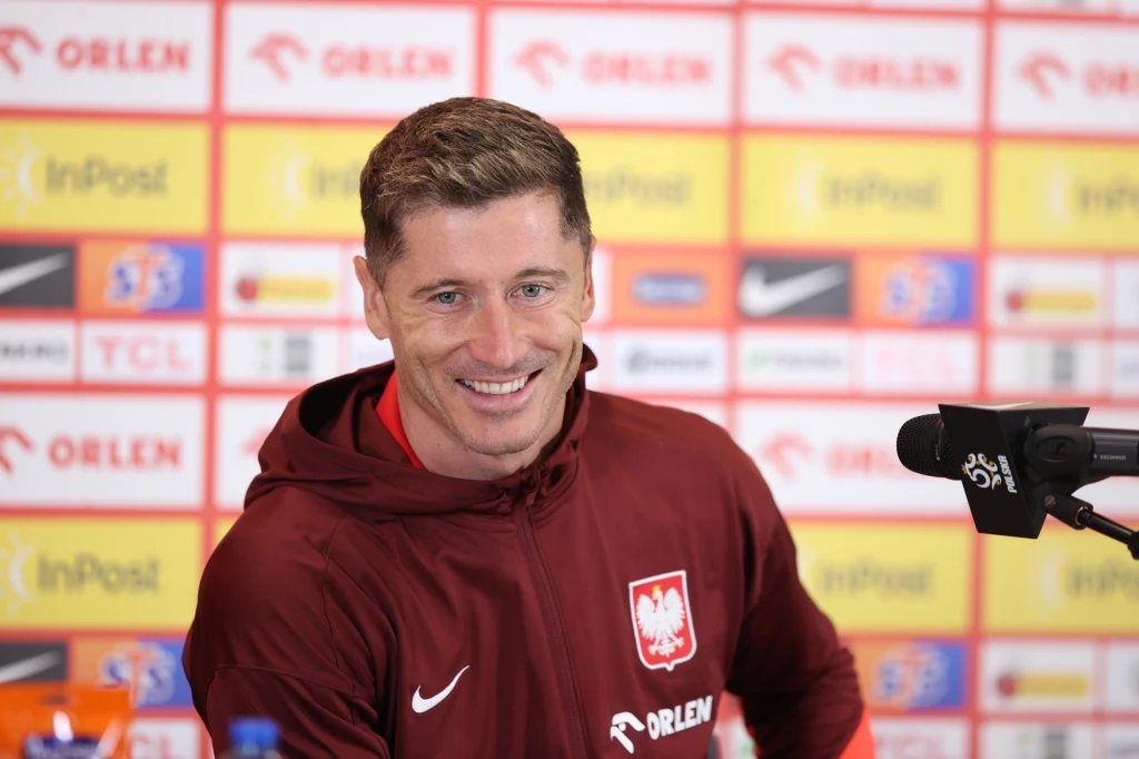 Robert Lewandowski Mężczyzna w sportowej bluzie reprezentacji Polski siedzi przy mikrofonie na tle ścianki sponsorskiej, uśmiecha się podczas konferencji prasowej.