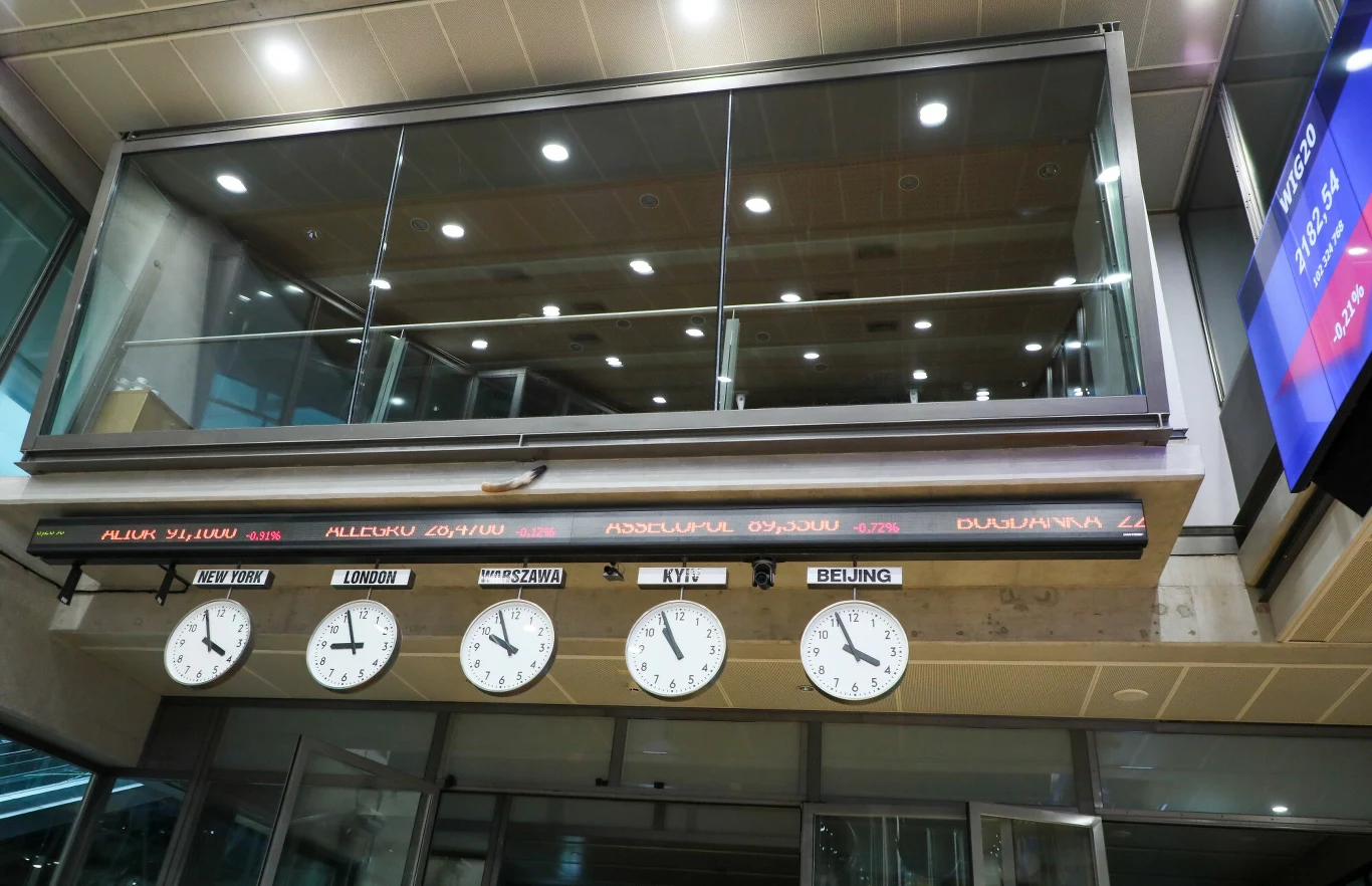 Dobre czasy nadeszły dla akcji na rynkach wschodzących. Od kilkunastu lat czegoś takiego nie widziano Pięć zegarów wyświetlających czas w różnych stolicach świata zawieszonych na ścianie pod cyfrową tablicą z nazwami miast i przesuwającymi się komunikatami, widoczne elementy nowoczesnego wnętrza budynku.