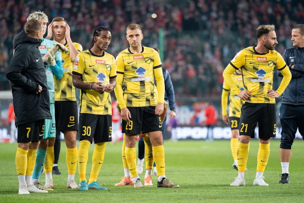 Grupa piłkarzy w żółto-czarnych strojach stoi na murawie stadionu, wokół widać rozmytą publiczność na trybunach, wyraz ich twarzy wskazuje na skupienie i analizę sytuacji po meczu.