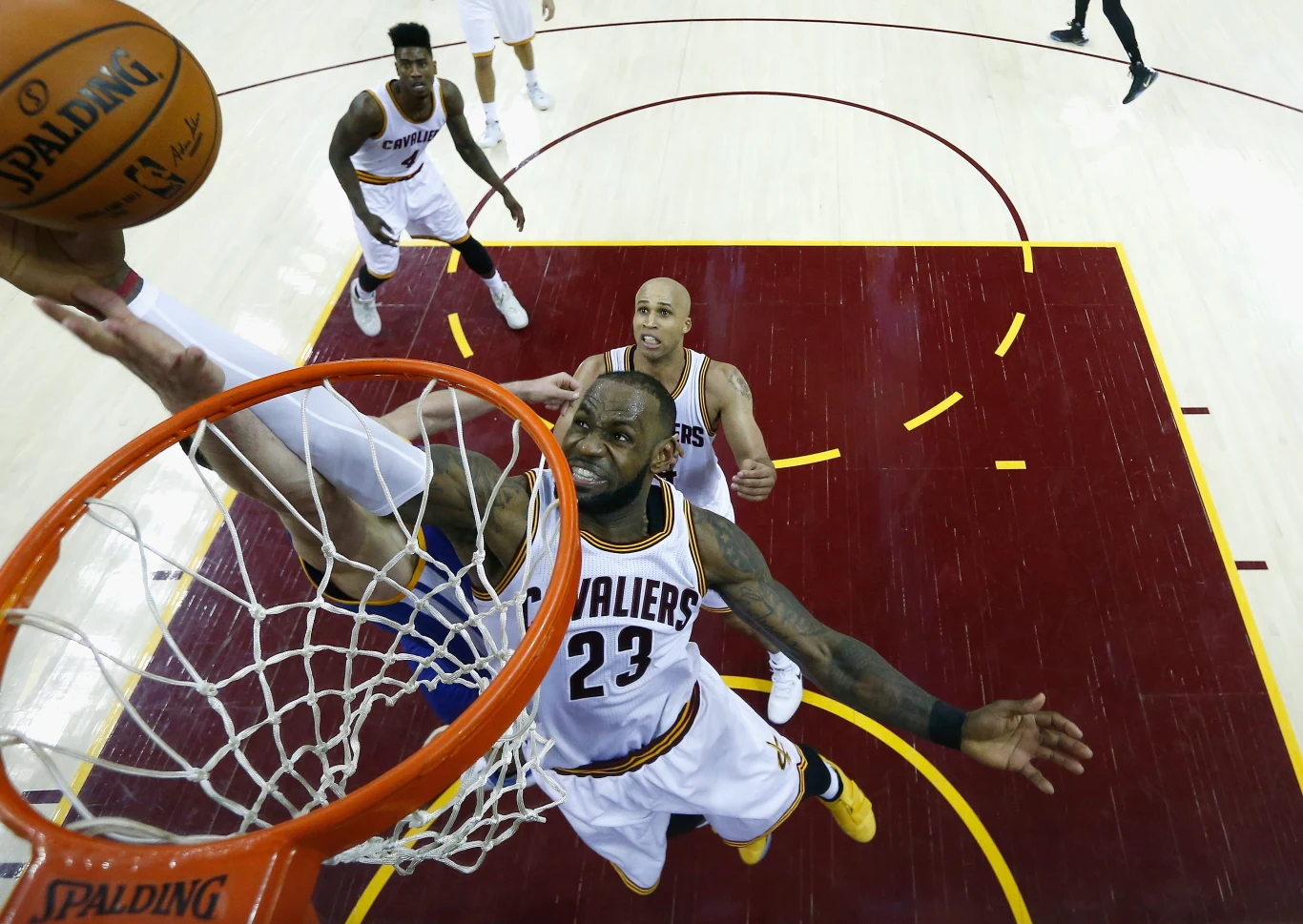 LeBron James w barwach Cleveland Cavaliers Koszykarz w białym stroju z numerem 23 próbuje zablokować zawodnika drużyny przeciwnej, który wykonuje wsad do kosza podczas meczu na profesjonalnym parkiecie. W tle widać kilku innych zawodników, obserwujących akcję.