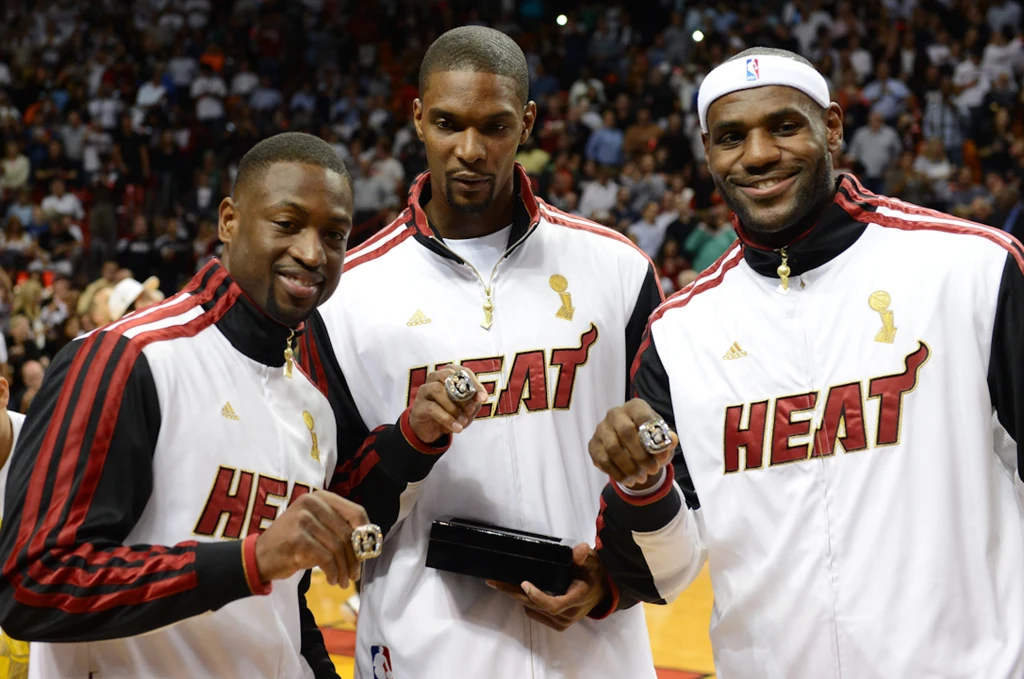 Dwyane Wade, Chris Bosh i LeBron James z pierścieniami za zdobycie mistrzostwa NBA z 2012 roku Trzech koszykarzy drużyny Miami Heat ubranych w klubowe dresy z mistrzowskimi pierścieniami na palcach prezentuje się na parkiecie przed licznie zgromadzoną publicznością; wszyscy uśmiechnięci i dumni ze zdobytego trofeum.