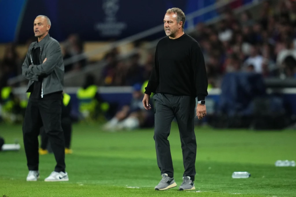 Hansi Flick Dwóch trenerów piłkarskich stoi na linii bocznej boiska podczas meczu, w tle rozmyta publiczność oraz ochrona w żółtych kamizelkach, atmosfera sportowego wydarzenia.