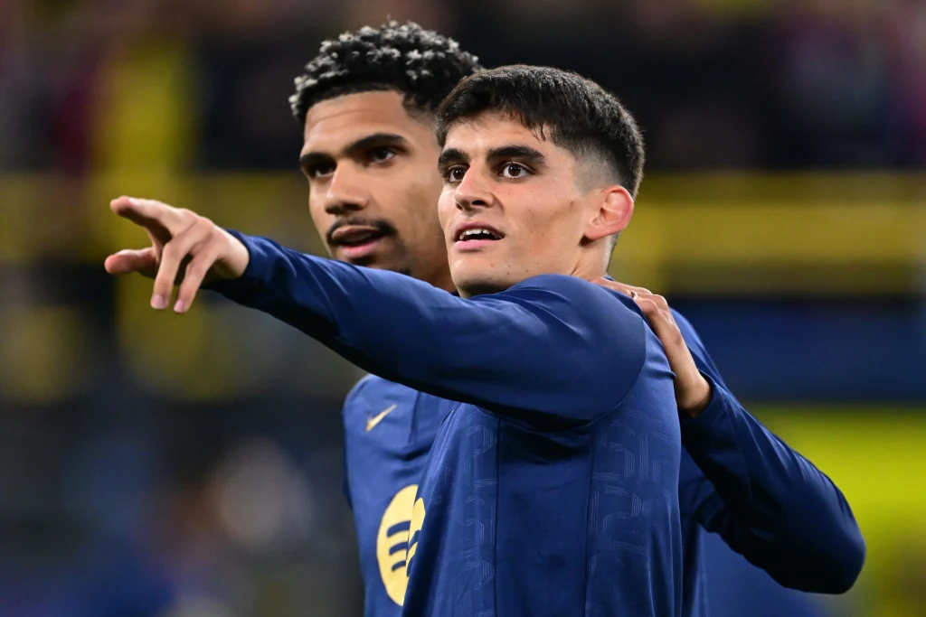 Ronald Araujo i Gerard Martin Dwóch młodych mężczyzn w sportowych strojach stoi blisko siebie, jeden z nich obejmuje drugiego ramieniem i wskazuje coś w oddali. Twarze są wyraźnie widoczne, tło rozmyte z elementami stadionu.