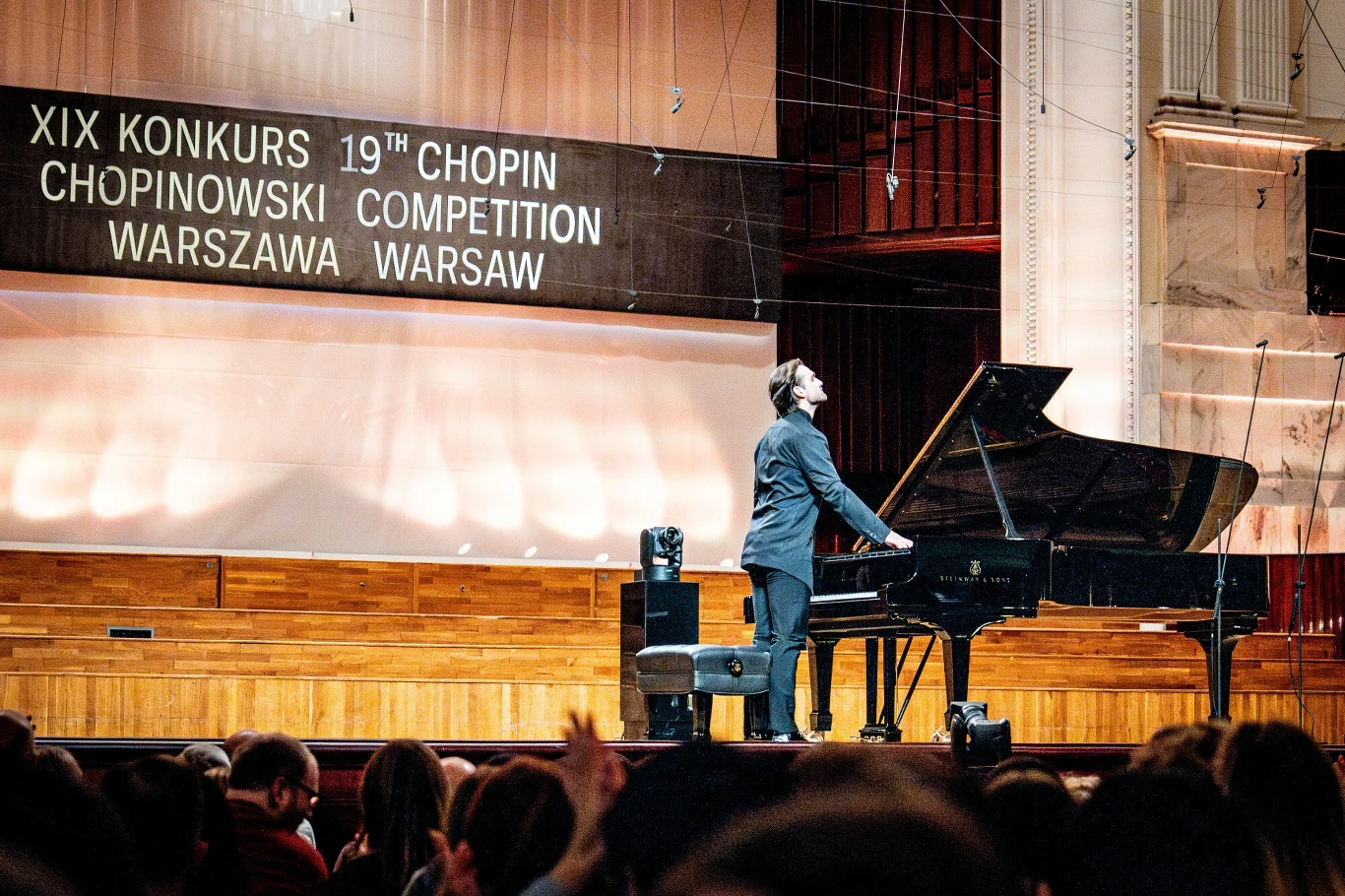 XIX Międzynarodowy Konkurs Pianistyczny im. Fryderyka Chopina w Warszawie Pianista w eleganckim stroju stojący przy fortepianie na scenie podczas XIX Konkursu Chopinowskiego w Warszawie, w tle widoczny duży baner informujący o wydarzeniu oraz skupiona publiczność siedząca na widowni.
