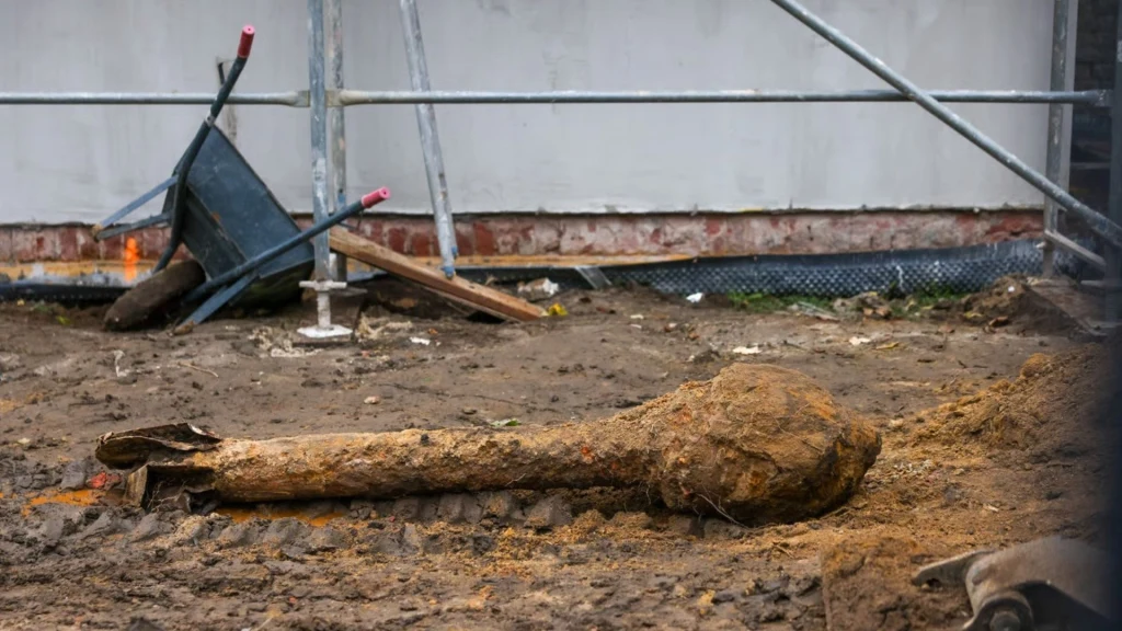 Duża, błotnista kość leży na ziemi na placu budowy, obok znajduje się przewrócona taczka oraz metalowe elementy rusztowania.