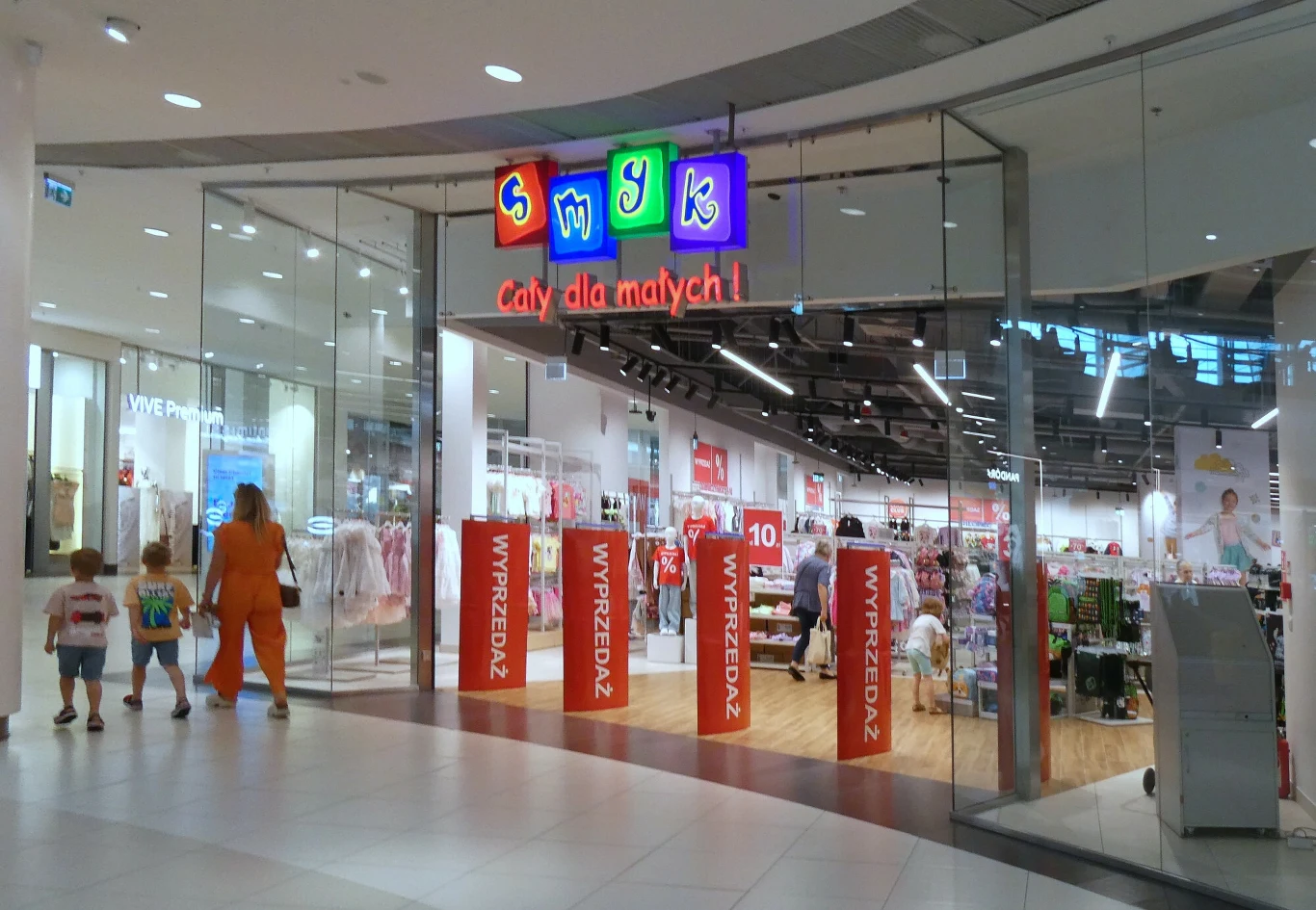 Sieć sklepów z artykułami dla dzieci Smyk planuje debiut na GPW Wejście do sklepu Smyk w centrum handlowym z kolorowym szyldem oraz napisem 'cały dla małych', widoczne promocje i wyprzedaże, kilka osób wchodzi do środka, wnętrze jasne i przestronne, dominują ubrania dziecięce.