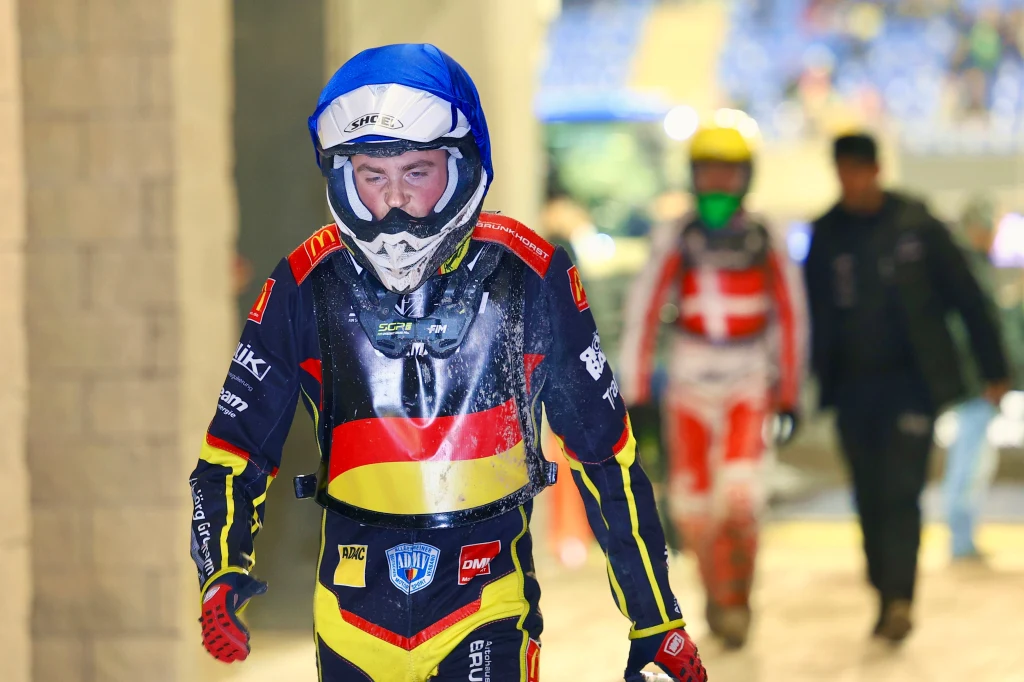 Norick Bloedorn podczas SoN2 w Toruniu Zawodnik żużlowy w pełnym kombinezonie i kasku po zakończonym wyścigu, na pierwszym planie, z widocznym zmęczeniem na twarzy; w tle inny zawodnik w stroju sportowym oraz rozmyte postacie.
