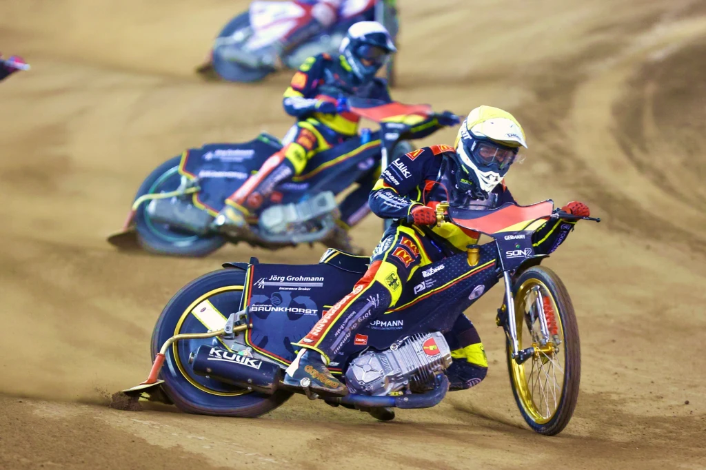 Norick Bloedorn Zawodnicy na motocyklach żużlowych rywalizują na łuku toru pokrytego nawierzchnią ziemną; motocykle odchylone, zawodnicy w kolorowych kombinezonach i kaskach, dynamiczna akcja wyścigu.