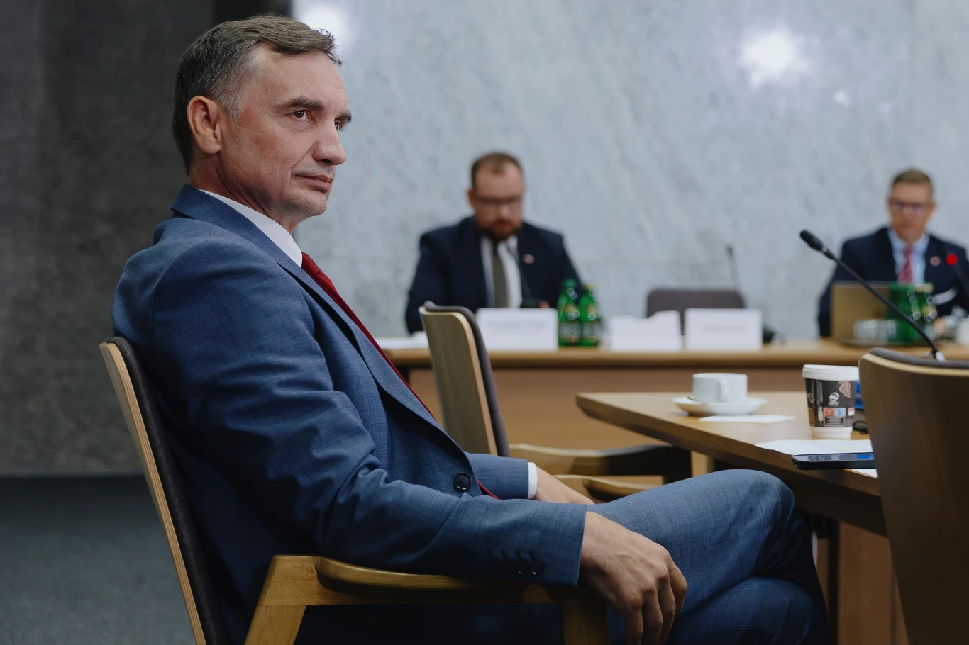 Komisja śledcza składa zawiadomienie na Zbigniewa Ziobrę Mężczyzna w niebieskim garniturze i czerwonym krawacie siedzący przy stole konferencyjnym, na drugim planie dwaj inni mężczyźni przy stole z mikrofonami, na stole kubek, filiżanka i butelki wody, tło z jasnym marmurem.