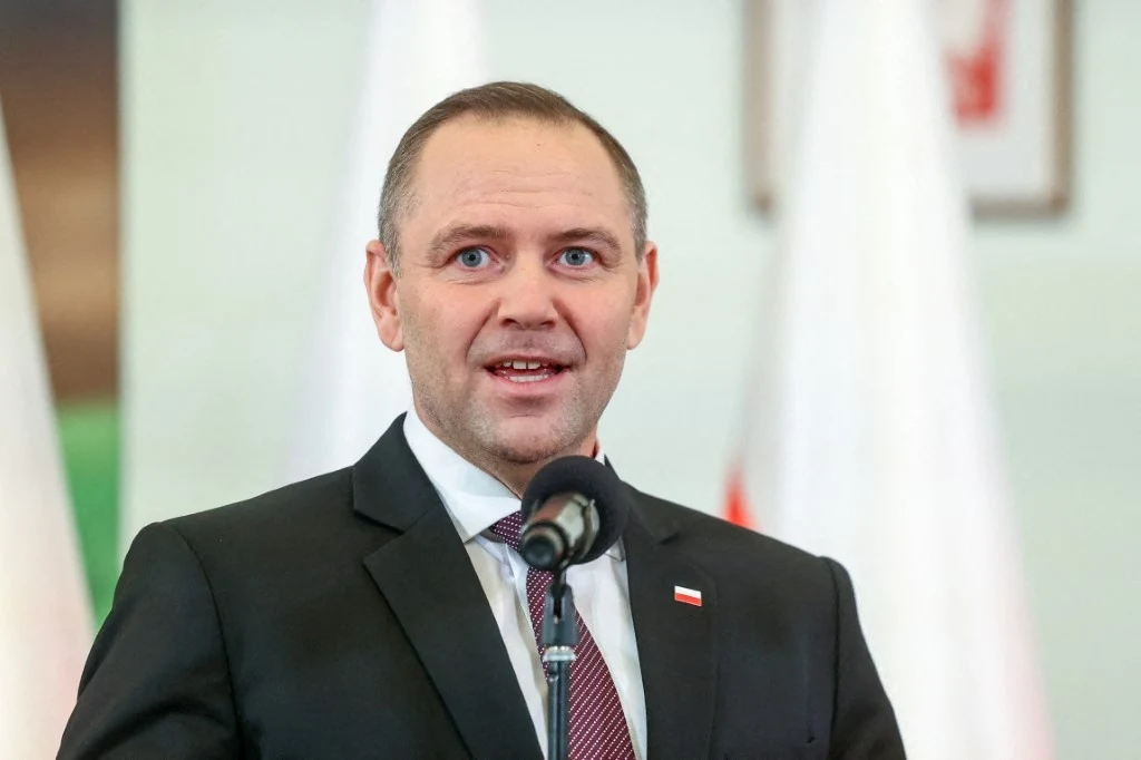 Karol Nawrocki Mężczyzna w garniturze i krawacie przemawia do mikrofonu, w tle widoczne biało-czerwone flagi, typowa oficjalna sceneria wystąpienia publicznego.