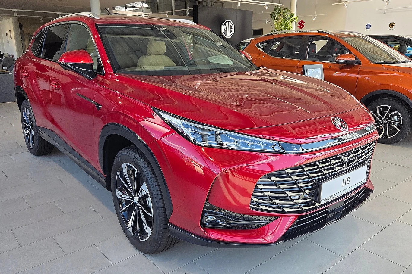 MG HS to pierwszy chiński samochód, który trafił na listę 10 najchętniej kupowanych nowych aut w Polsce Czerwony samochód typu SUV marki MG stoi zaparkowany w nowoczesnym salonie samochodowym, z widocznymi innymi modelami aut w tle, jasnym oświetleniem i eleganckim wykończeniem wnętrza.