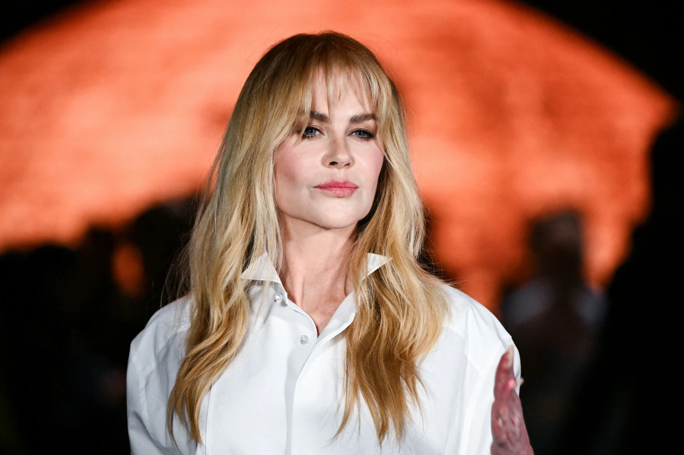 Nicole Kidman na Paryskim Tygodniu Mody Kobieta z długimi blond włosami i jasną koszulą patrzy prosto w obiektyw, w tle rozmyte pomarańczowe światło przyciąga uwagę i tworzy kontrastową atmosferę.