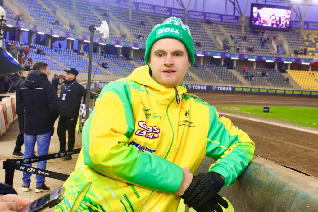 Mężczyzna w kurtce sportowej w kolorach żółtym i zielonym oraz zielonej czapce stoi przy bandzie stadionu żużlowego, w tle widoczne trybuny i fragment toru.