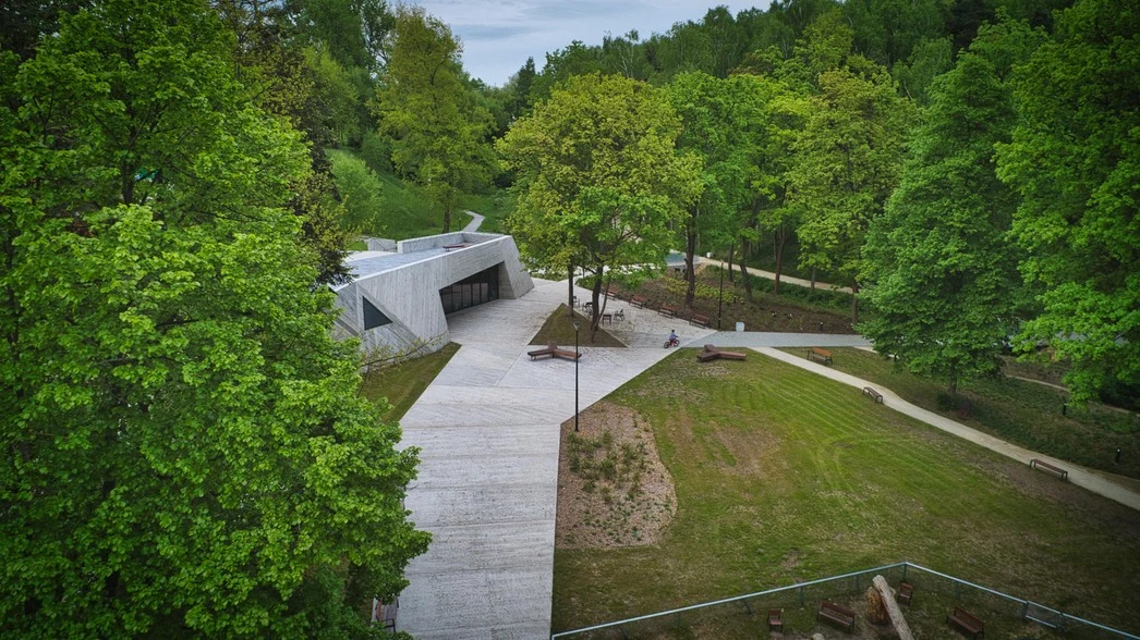 Nowoczesny budynek o geometrycznych kształtach otoczony zielonym parkiem oraz alejkami spacerowymi, w tle gęsty las liściasty, jasne chodniki i uporządkowana przestrzeń terenowa.