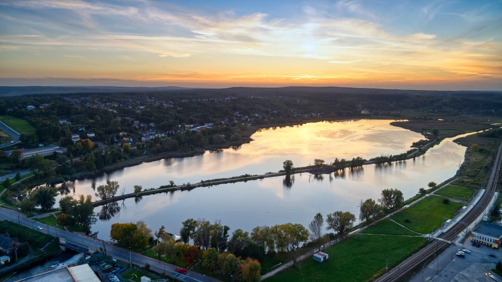 Starachowice z lotu ptaka fot. UM Starachowice Duże jezioro otoczone drzewami oraz zabudowaniami, widoczne przy zachodzie słońca, tafla wody odbija kolorowe niebo, w tle niewielkie wzgórza i zabudowania miejskie.