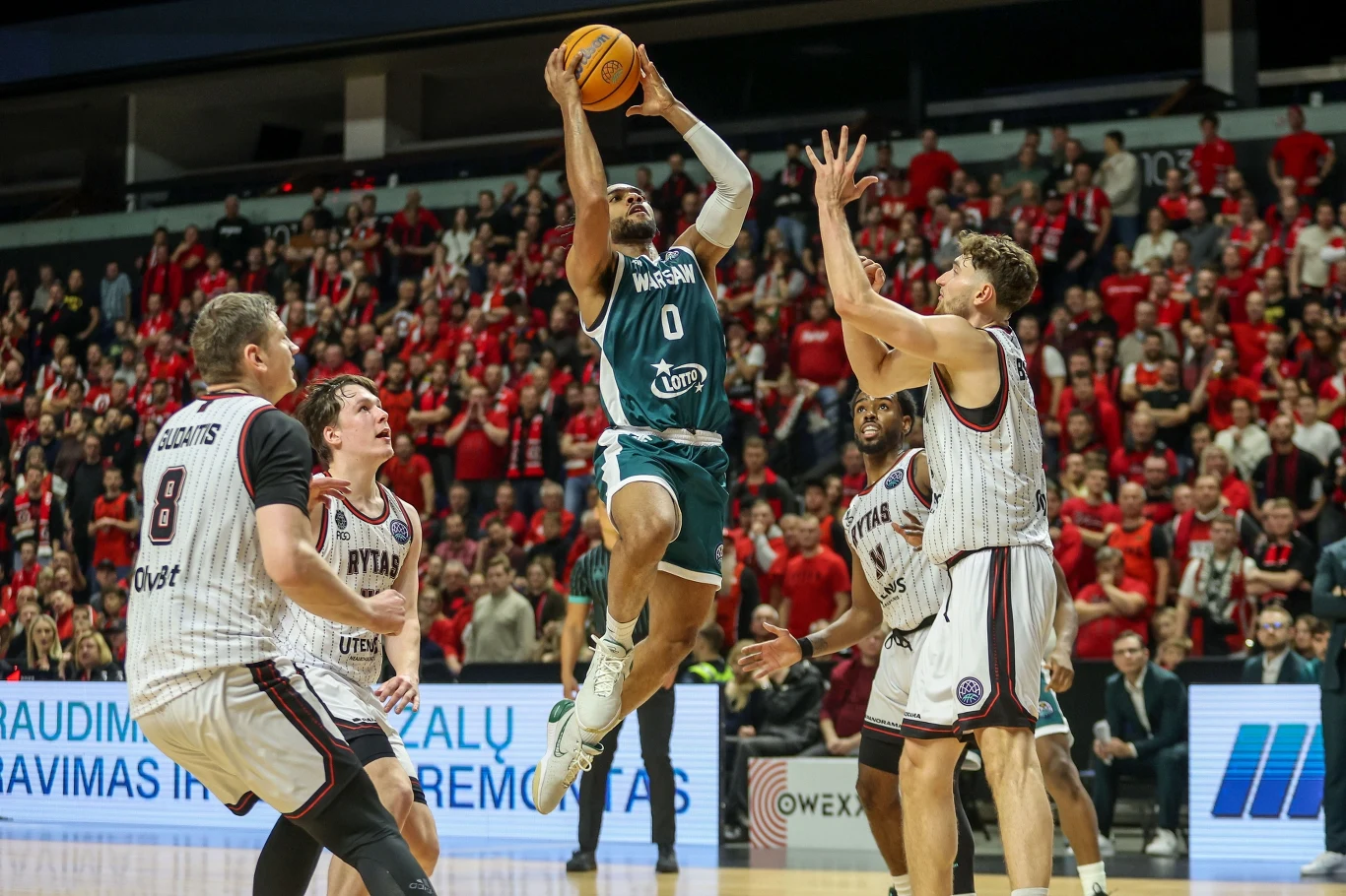 Mecz Rytas - Legia Koszykarz w zielonym stroju wyskakuje pod koszem z piłką do rzutu, otoczony przez trzech zawodników drużyny przeciwnej w biało-czerwonych strojach na tle licznej publiczności ubranej w czerwone koszulki.