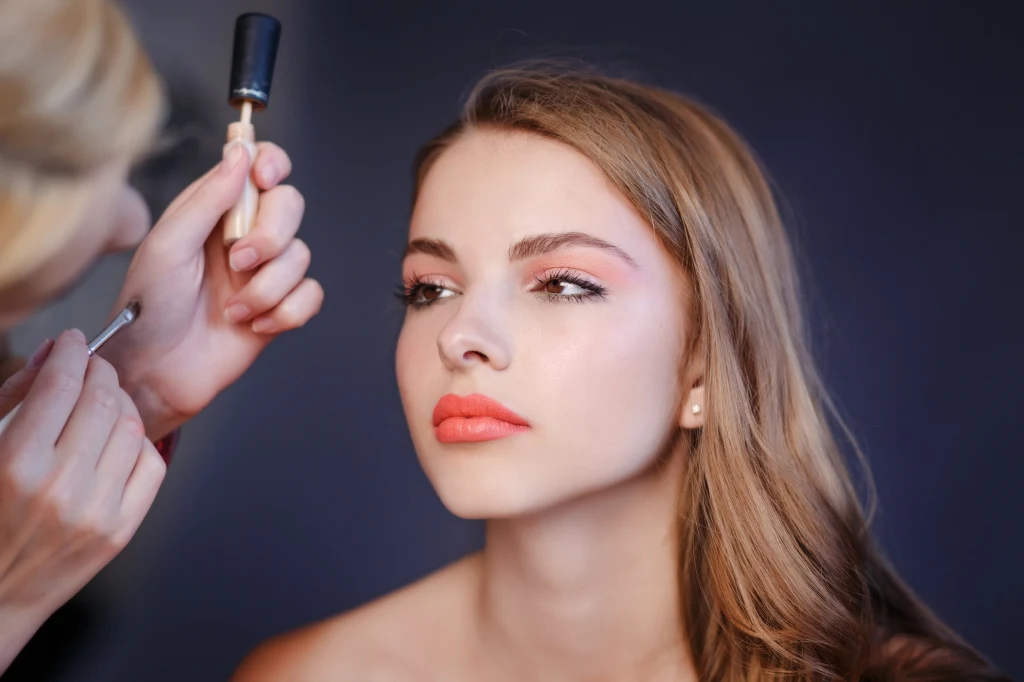 Kolory szminek i cieni mogą rozświetlić jesienną cerę Młoda kobieta z długimi blond włosami siedzi, a wizażystka aplikuje jej makijaż, skupiając się na jej oczach, twarz jest delikatnie podkreślona, a usta pomalowane na intensywny kolor