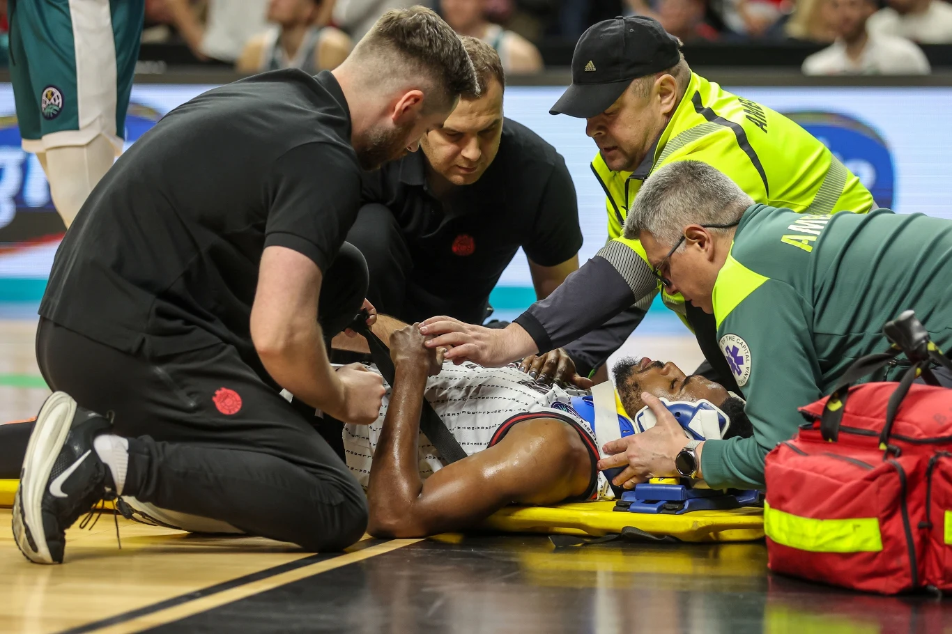 Jerrick Harding otrzymujący pomoc medyczną Koszykarz leżący na plecach na boisku podczas meczu otaczany przez czterech ratowników udzielających pomocy medycznej. Zawodnik jest wyraźnie poszkodowany, a ratownicy wyposażeni są w medyczne torby i sprzęt.