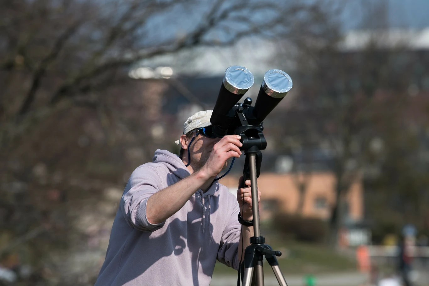 Mężczyzna w jasnej bluzie i czapce obserwuje niebo przez teleskop na statywie, sytuacja odbywa się na zewnątrz w słoneczny dzień, w tle rozmyty krajobraz miejski.