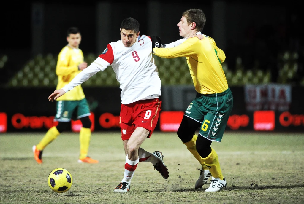 Robert Lewandowski podczas meczu z Litwą w 2011 roku dwóch piłkarzy walczy o piłkę podczas meczu, zawodnik w biało-czerwonym stroju jest blokowany przez gracza w żółto-zielonym stroju, w tle inni zawodnicy oraz rozmyta publiczność