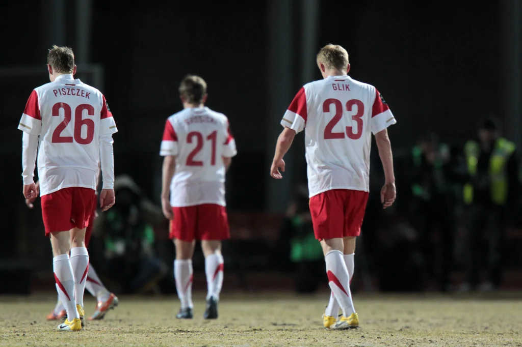 Łukasz Piszczek i Kamil Glik podczas meczu z Litwą w 2011 roku Trzech piłkarzy w biało-czerwonych strojach z nazwiskami Piszczek, Glik i numerami na plecach schodzi z boiska na murawie, w oddali widoczna grupa fotoreporterów oraz zaciemnione tło stadionu.